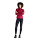 En kvinde med kort, krøllet hår står med ansigtet vendt fremad og armene over kors. Hun er iført en rød langærmet sportstrøje, sorte New Balance Athletics Heat High Rise Legging 27″ med NB HEAT-teknologi samt sorte sneakers, alt sammen på en ensfarvet hvid baggrund.
