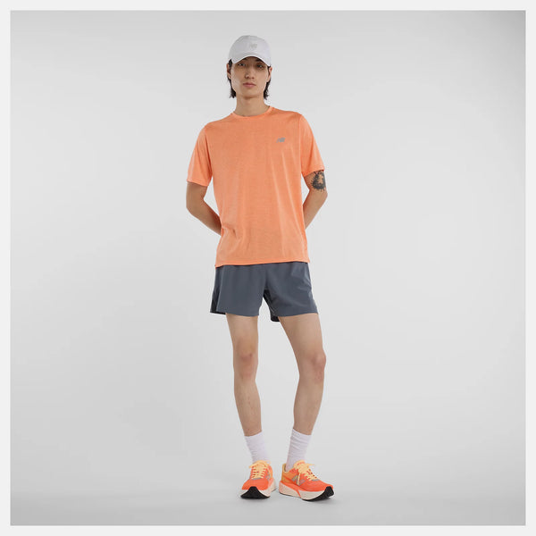 En person står på en hvid baggrund iført New Balance Athletics T-shirt, Herre, Aae med hurtigtørrende teknologi, grå sportsshorts, en lysegrå kasket, hvide sokker og orange løbesko. Den ene arm hviler på hoften.