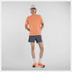 En person står på en hvid baggrund iført New Balance Athletics T-shirt, Herre, Aae med hurtigtørrende teknologi, grå sportsshorts, en lysegrå kasket, hvide sokker og orange løbesko. Den ene arm hviler på hoften.