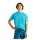 En person med mellemlangt brunt hår har en lyseblå New Balance Athletics T-shirt (Herre, Altitude) og mørkeblå shorts på og står foran en ensfarvet hvid baggrund med den ene hånd på hoften.