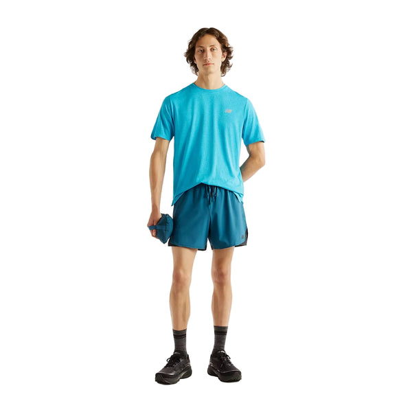 En mand står med ansigtet vendt fremad iført en turkis New Balance Athletics-T-shirt, Herre, Altitude og blå shorts samt grå sokker og sorte sneakers. Han holder en lille blå taske i hånden; den ensfarvede hvide baggrund fremhæver hans look.