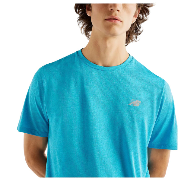 En person er iført en lyseblå New Balance Athletics-T-shirt, Herre, Altitude, foran en hvid baggrund. Den øverste del af overkroppen og en del af ansigtet er synlige, ligesom T-shirtens logo på venstre bryst.