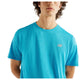 En person er iført en lyseblå New Balance Athletics-T-shirt, Herre, Altitude, foran en hvid baggrund. Den øverste del af overkroppen og en del af ansigtet er synlige, ligesom T-shirtens logo på venstre bryst.