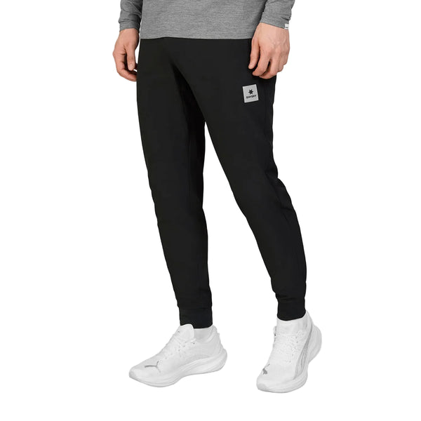 En person iført en grå langærmet trøje, Saysky Blaze-bukser (unisex, sorte) med et lille logo-mærke og hvide sportssko står foran en ensfarvet hvid baggrund.