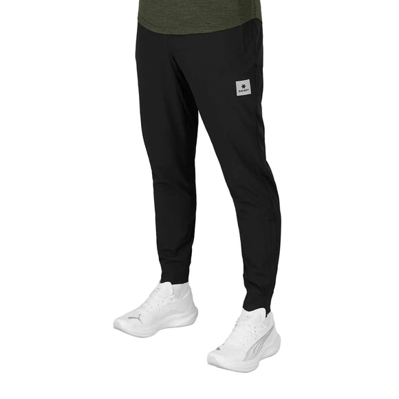 Man ser en person fra livet og ned, iført Blaze Pants, Unisex, Sort fra Saysky, med et gråt mærke på venstre ben og hvide sportssko. Den ensfarvede hvide baggrund fremhæver disse stilfulde træningsbukser.