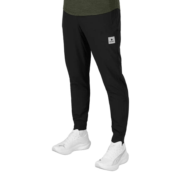 Man ser en person fra livet og ned, iført Blaze Pants, Unisex, Sort fra Saysky, med et gråt mærke på venstre ben og hvide sportssko. Den ensfarvede hvide baggrund fremhæver disse stilfulde træningsbukser.