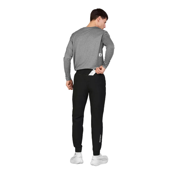 En mand iført en grå langærmet skjorte og sorte Blaze-bukser fra Saysky (unisex) står med ryggen til og lægger en hvid genstand i baglommen på sine bukser. Baggrunden er helt hvid.