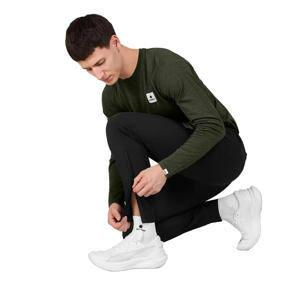 En mand iført en grøn langærmet skjorte og et par Saysky Blaze-bukser, unisex, sorte, sidder på hug, mens han åbner lynlåsen i underbenet på bukserne. Han har desuden hvide sportssko og hvide sokker på. Baggrunden er ensfarvet og lys.