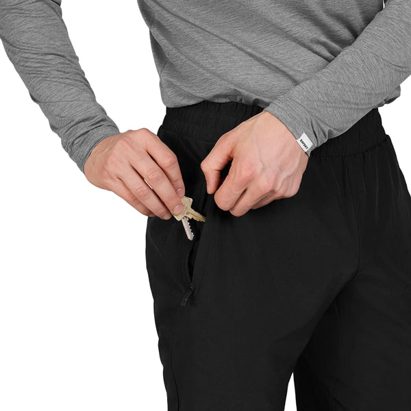 Der ses en person iført en grå langærmet trøje og sorte bukser – nærmere bestemt Saysky Blaze Pants, Unisex, Black – der putter en nøgle i lommen med lynlås på disse træningsbukser. Personens ansigt kan ikke ses.