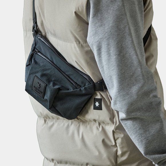 En person i en lys beige pufferjakke med et gråt ærme ses fra siden og bærer Saysky Bum Bag 601, Unisex, Black med en stjernelogo-patch og lynlåslommer.