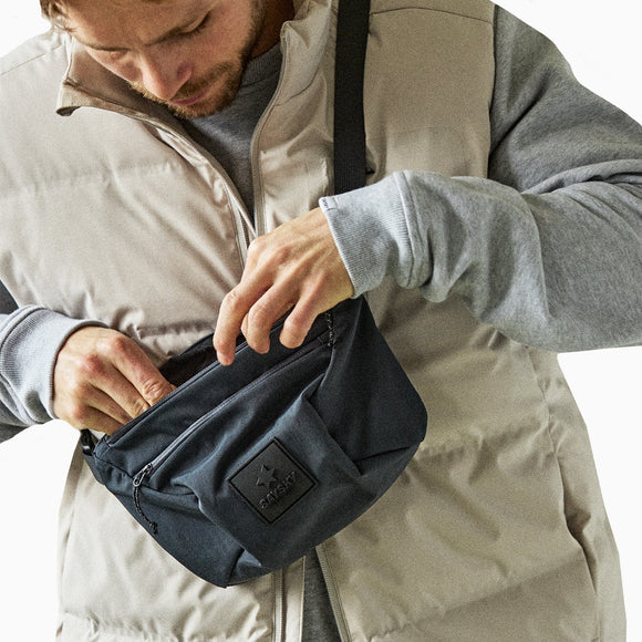 En person i en lys puffer-vest og grå sweatshirt kigger ned, mens han rækker ned i en sort Saysky Bum Bag 601 (unisex) med en patch foran.