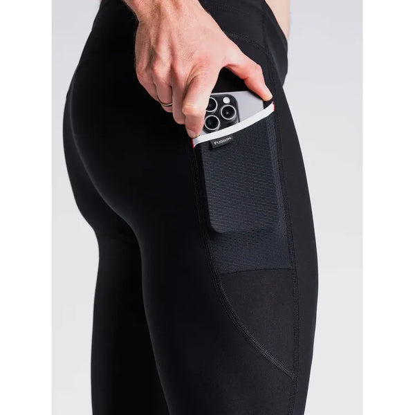 En person iført Fusion Long Tight Unisex SuperBlack leggings lægger en smartphone i sidelommen, mens billedet fremhæver lommen og hånden, der holder telefonen.