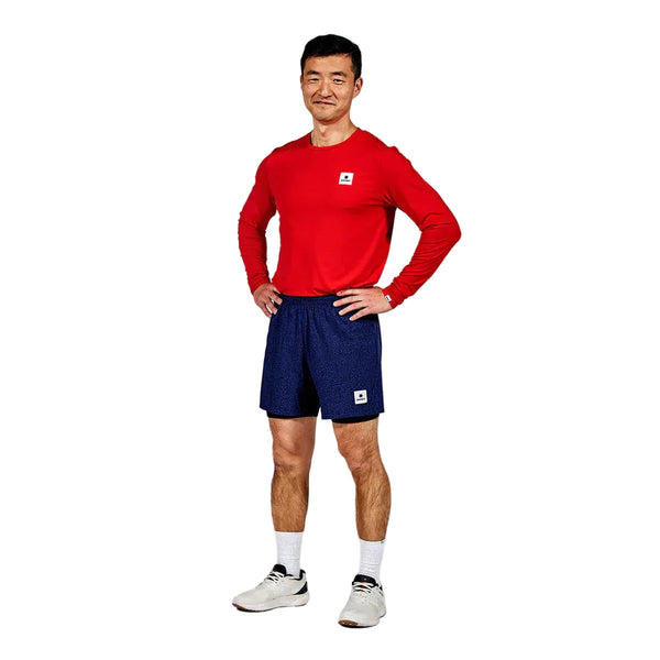 En mand står med ansigtet fremad og hænderne på hofterne, iført en rød langærmet skjorte, Saysky CC Pace 2 in 1 Shorts 5" Herre i blå - ideel til maratonløbere - samt hvide sokker og sneakers, på en almindelig hvid baggrund.