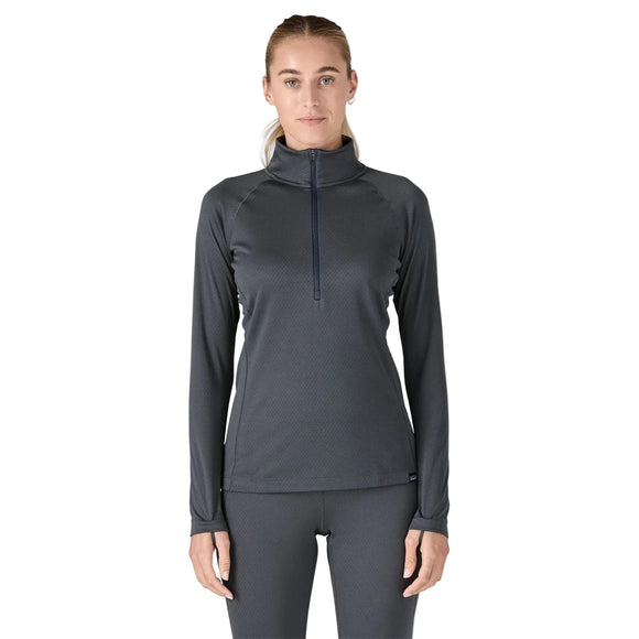 En kvinde står med ansigtet fremad og er iført en Patagonia Cap Midweight Zip Neck til kvinder i Smolder Blue med matchende leggings. Hendes hår er trukket tilbage, og hendes arme hviler ved siden af hende mod en almindelig hvid baggrund.
