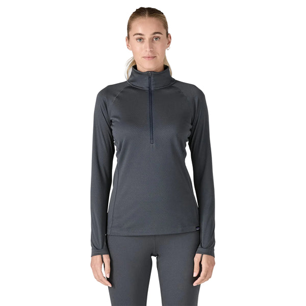 En kvinde står med ansigtet fremad og er iført en Patagonia Cap Midweight Zip Neck til kvinder i Smolder Blue med matchende leggings. Hendes hår er trukket tilbage, og hendes arme hviler ved siden af hende mod en almindelig hvid baggrund.