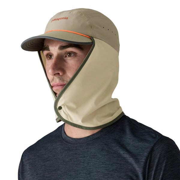 En mand bærer unisex Patagonia Caped Merganzer Hat i River Rock Green med en beige kasket, grøn kant og et påsat ventileret ansigts- og halsdæksel. Han står i en mørkeblå skjorte og kigger lidt til venstre mod en hvid baggrund.
