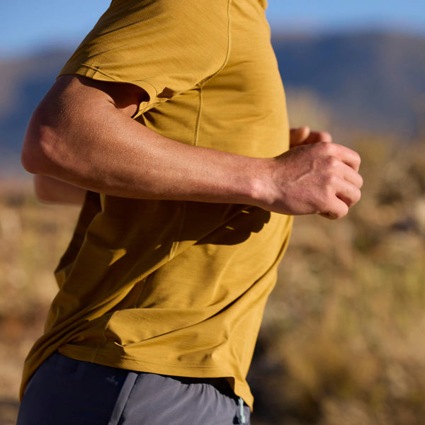 En mand iført en Patagonia Capilene Cool Ultra Shirt i Bobcat Brown - Light Bobcat Brown X-Dye og grå shorts løber udendørs med tørt græs og slørede bjerge bag sig. Kun hans overkrop er synlig.