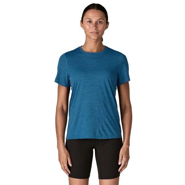 En person står med ansigtet fremad i en Patagonia Capilene Cool Ultra Shirt, Dame, Aquatic Blue - Light Aquatic Blue X-Dye - en åndbar løbetrøje - sammen med sorte shorts på en almindelig hvid baggrund.