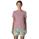 En kvinde med glat, brunt hår, der er trukket tilbage, bærer en Patagonia Capilene Cool Ultra Shirt, Dame, i Light Violet - Quiet Violet X-Dye og flerfarvede, mønstrede shorts. Hun står med ansigtet fremad mod en almindelig hvid baggrund.