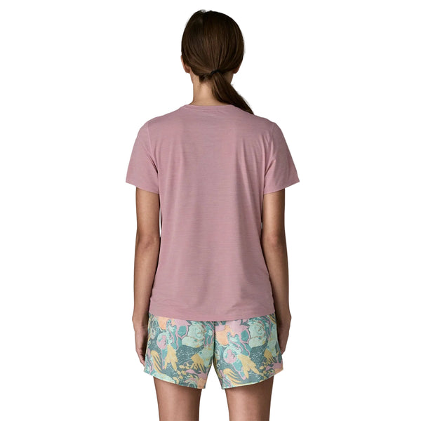 En person med langt brunt hår i en lav hestehale står med ryggen til kameraet iført Patagonia Capilene Cool Ultra Shirt til kvinder i Light Violet - Quiet Violet X-Dye og pastelfarvede shorts med tryk.