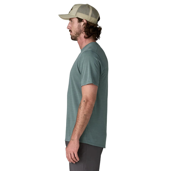 En mand står i profil mod venstre iført Patagonias Capilene Cool Ultra Shirt til mænd i Blue Sage - Light Blue Sage X-Dye, sammen med mørkegrå shorts og en beige truckerhat. Den ensfarvede hvide baggrund fremhæver skjortens stil og komfort.