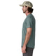 En mand står i profil mod venstre iført Patagonias Capilene Cool Ultra Shirt til mænd i Blue Sage - Light Blue Sage X-Dye, sammen med mørkegrå shorts og en beige truckerhat. Den ensfarvede hvide baggrund fremhæver skjortens stil og komfort.
