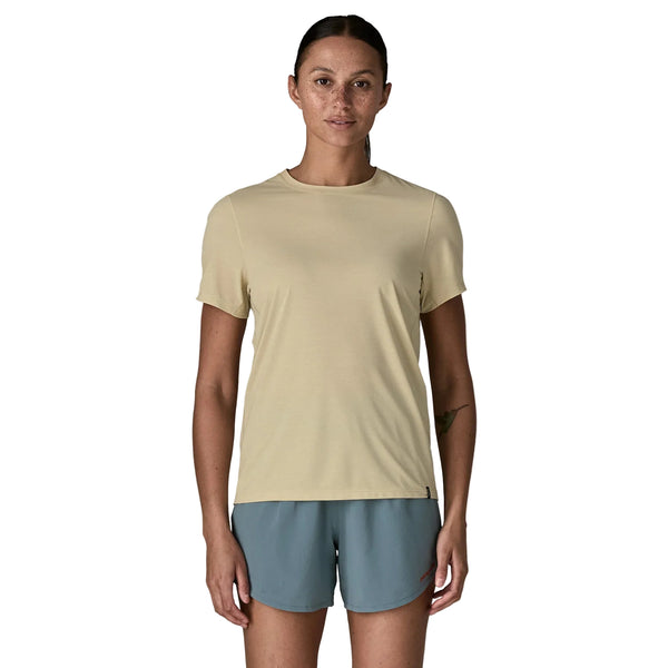 En person med mørkt hår, der er bundet tilbage, er iført Patagonia Capilene Cool Ultra Shirt, Dame, Pumice - Dyno White X-Dye og blågrå shorts og står med front mod en almindelig hvid baggrund.