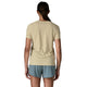 En kvinde med langt mørkt hår i hestehale står med ansigtet vendt væk og er iført Patagonia Capilene Cool Ultra Shirt Dame (Pumice - Dyno White X-Dye) og lyseblå shorts. En lille tatovering er synlig nær hendes højre albue. Baggrunden er almindelig hvid.