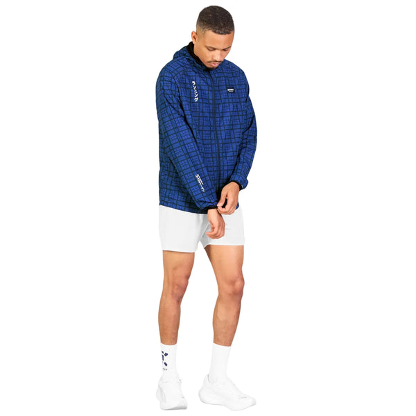 En mand står foran en hvid baggrund iført Saysky Checker Pace-jakken, Herre, Blue Checker, kombineret med hvide shorts, hvide sokker med blå detaljer og hvide sneakers. Han kigger ned, mens han med den ene hånd retter ærmet på sin Saysky-løbejakke, hvilket understreger både komfort og vindbeskyttelse.