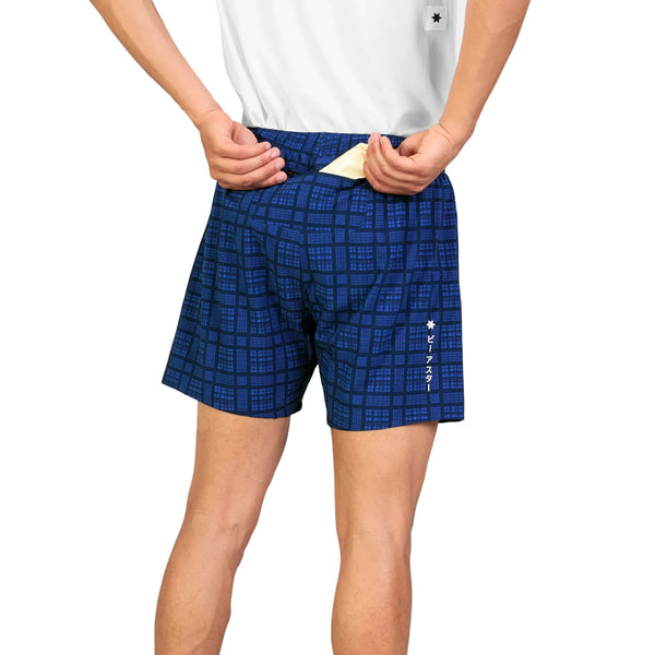 En person iført en hvid skjorte og Saysky Checker Pace Shorts 5″ (Herre, Blue Checker) tager en gul genstand frem fra baglommen. På venstre side af træningsshortsene ses hvide symboler og japanske tegn. Personen ses bagfra, fra taljen til midt på låret.