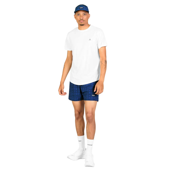 En mand står foran en hvid baggrund iført en hvid T-shirt, Saysky Checker Pace Shorts 5″ i Blue Checker, hvide strømper, hvide sneakers og en blå kasket. Dette klassiske herrelook forener afslappet stil og atletisk funktionalitet med højtydende shorts designet af Saysky, der sikrer komfort hele dagen. Han står med ansigtet vendt fremad, venstre arm afslappet ned langs siden og højre arm let bøjet.