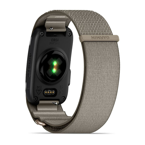 Et Garmin Cirqa Smart Band i farven French Grey vises set bagfra, hvor dets stofrem og det synlige grønne sensorlys under urskiven fremhæves. Som et alsidigt aktivitetsarmbånd byder det på funktioner såsom søvnregistrering. Remmen er afsluttet med en lås, der er forsynet med et Garmin-mærke.