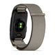 Et Garmin Cirqa Smart Band i farven French Grey vises set bagfra, hvor dets stofrem og det synlige grønne sensorlys under urskiven fremhæves. Som et alsidigt aktivitetsarmbånd byder det på funktioner såsom søvnregistrering. Remmen er afsluttet med en lås, der er forsynet med et Garmin-mærke.