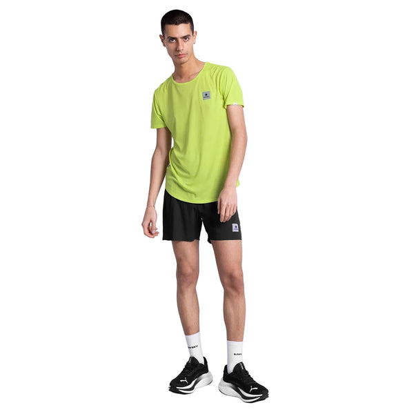 En person står på en hvid baggrund iført en Saysky Reactive Flow T-shirt, Herre, Yellow Logo, sammen med sorte shorts, hvide sokker og sorte sneakers. Hovedet er let på skrå, og den ene arm hænger løst ned langs siden.