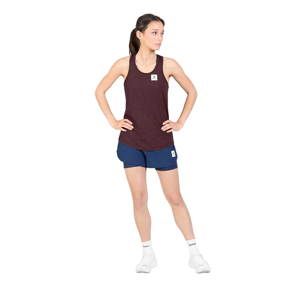 En person står foran en hvid baggrund iført Saysky W Clean Combat Pro Singlet, Dame, i farven burgundy, fremstillet af DELTAPEAK Carat-stof, kombineret med blå sportsshorts, hvide sokker og hvide sneakers. Med hænderne på hofterne og blikket vendt mod siden er dette hurtigtørrende sæt ideelt til aktive dage.