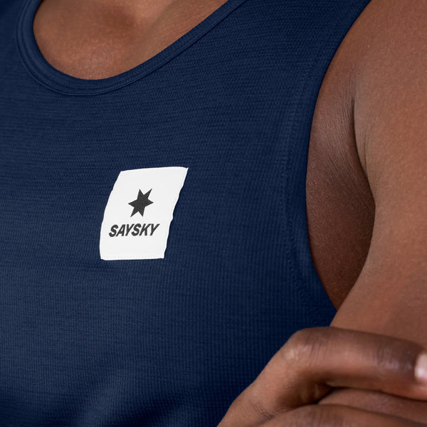 En person har en Saysky Clean Combat Pro Singlet, Herre, Blå, på, der er fremstillet af DELTAPEAK-stof og har et hvidt mærke med en sort stjerne og teksten »SAYSKY« trykt på brystet. Personens hånd kan ses i nederste højre hjørne.