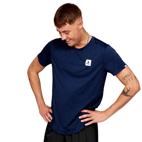 En mand med kort brunt hår og synlige tatoveringer på armene bærer en marineblå Saysky Clean Combat Pro T-shirt, Herre, Blue med et lille hvidt logo på brystet. Han står med hænderne på hofterne, kigger ned og smiler.
