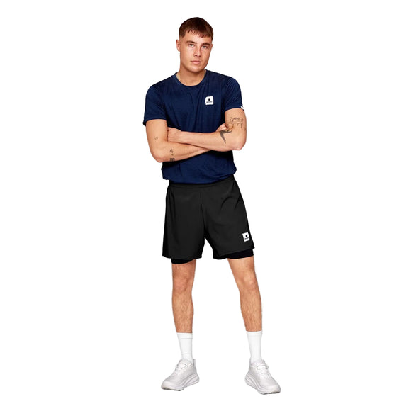 En person står med armene over kors iført en Saysky Clean Combat Pro T-shirt, Herre, Blue, sammen med sorte shorts, hvide sokker og hvide sportssko på en almindelig hvid baggrund.