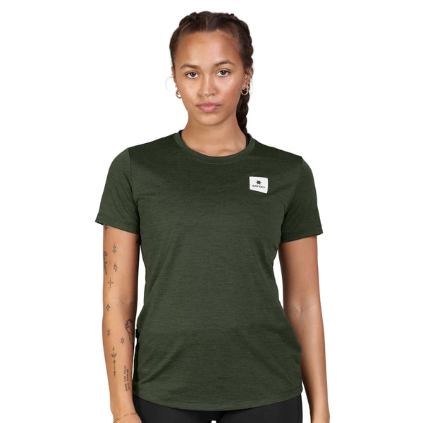 En person med flettet hår har en Saysky Clean Combat Pro T-shirt, Dame, i grøn på, med et lille hvidt logo-mærke på venstre bryst. Personen har synlige tatoveringer på armene og står foran en ensfarvet hvid baggrund.