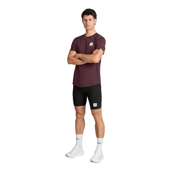 En person står med ansigtet fremad og armene over kors, iført en Saysky Clean Combat Pro T-shirt, Herre, Burgundy, kombineret med sorte shorts, hvide strømper og hvide sneakers. Den åndbare løbetrøje fra Saysky er både let og hurtigtørrende, hvilket gør den ideel til aktiv brug. Baggrunden er ensfarvet hvid.