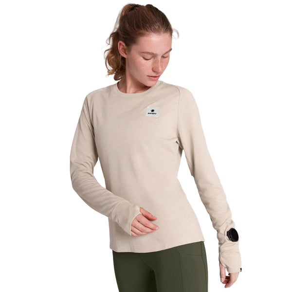 En kvinde har en beige Saysky Clean Flow langærmet sportstrøje på med tommelfingerhuller og et lille logo-mærke på brystet.
