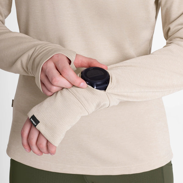 En person, der har en beige Clean Flow-langærmet trøje med manchetter med tommelfingerhuller på, trækker ærmet op for at se på sit smartwatch på håndleddet.