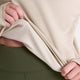 Hænderne løfter kanten af en beige Clean Flow-langærmet bluse og afslører det strukturerede for og de olivengrønne leggings nedenunder.