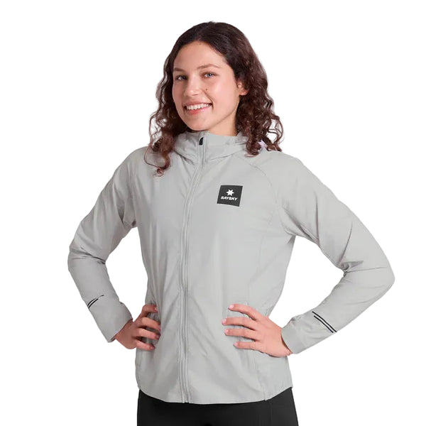 En kvinde med langt krøllet hår smiler, mens hun har Saysky Clean Pace Flex Hooded Jacket, Dame, Grey på - en lysegrå vindtæt løbejakke med et lille sort og hvidt logo på brystet og sorte lynlåse på ærmerne. Baggrunden er hvid.