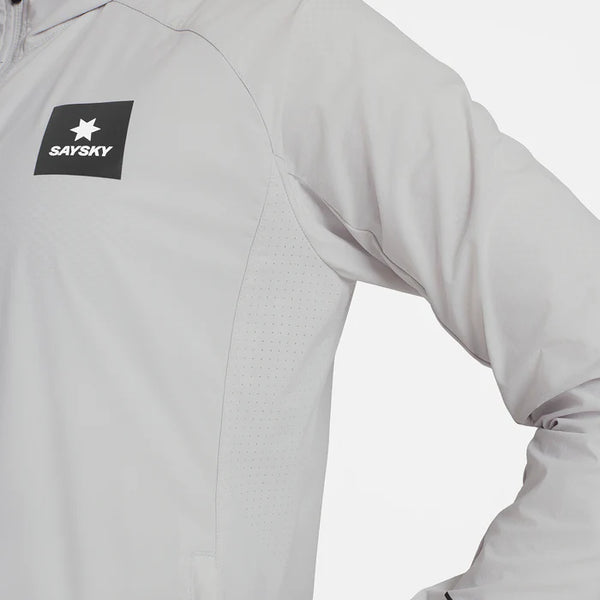 Nærbillede af en person iført den lysegrå Saysky Clean Pace Flex Hooded Jacket, Dame, med et sort mærke med en hvid stjerne og SAYSKY-logo på venstre bryst; deres højre hånd hviler på hoften.