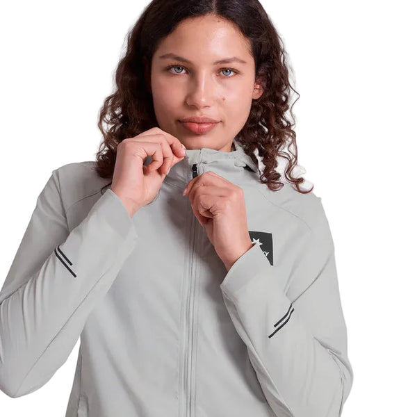 En kvinde med krøllet hår ses med taljen i vejret, mens hun lyner en lysegrå Saysky Clean Pace Flex Hooded Jacket, Dame, Grey. Den hvide baggrund fremhæver denne stilfulde og vindtætte løbejakke, som er perfekt til aktive kvinder på farten.