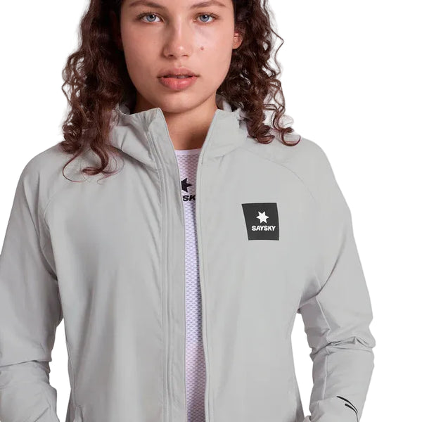 En kvinde med krøllet brunt hår bærer Saysky Clean Pace Flex Hooded Jacket, Dame, Grey. Den vindtætte løbejakke har lynlås, hætte og logo på brystet. Hun står på en almindelig hvid baggrund.