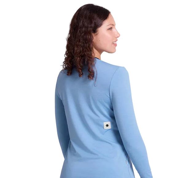En kvinde med krøllet brunt hår kigger væk iført Saysky Clean Pace Long Sleeve, Dame, Blue - en hurtigtørrende skjorte med en sort stjerne på ryggen. Den ensfarvede hvide baggrund fremhæver den løbetidsklare stil.