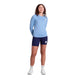En kvinde kigger fremad med hænderne på ryggen, iført Saysky Clean Pace Long Sleeve (Dame, Blue), mørkeblå shorts, hvide sokker og hvide sko - ideelt til løbeture - mod en almindelig hvid baggrund.