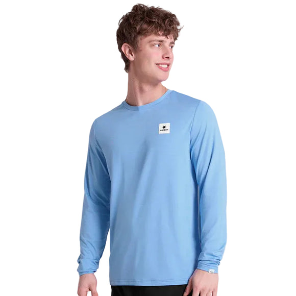 En ung mand med kort krøllet hår bærer Saysky Clean Pace Long Sleeve, Herre, Blue, med et lille firkantet hvidt mærke på brystet. Han står på en almindelig hvid baggrund, kigger til venstre og smiler lidt.